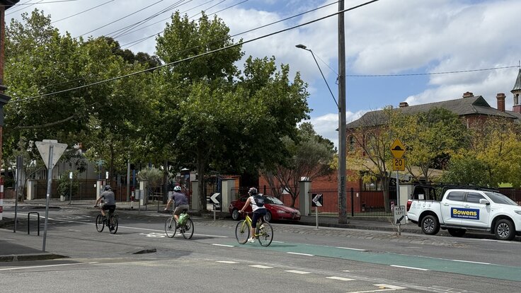 Three people on bikes on the green bike lane on Wellington Street in Clifton Hill, passing the school