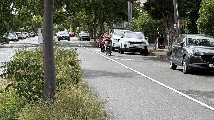 A parent with a child in the front seat of a cargo e-bike rides along Wellington Street between parked cars and moving traffic, with no protected cycling lane